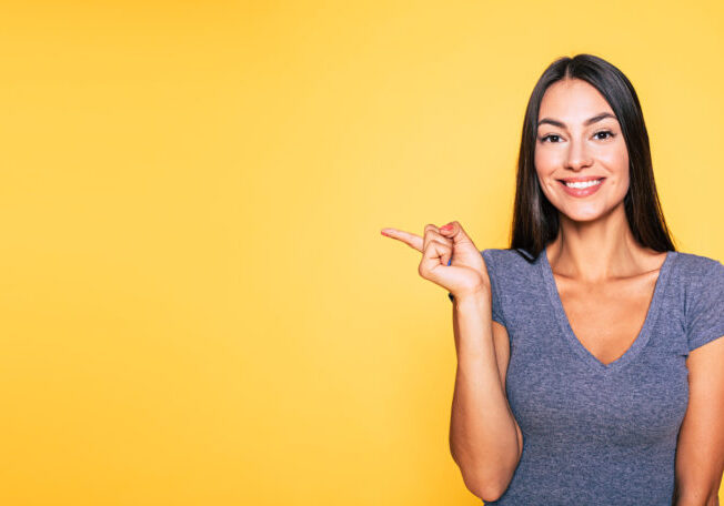 Photo of young excited beautiful happy brunette woman, girl pointing away and smile isolated on yellow background banner